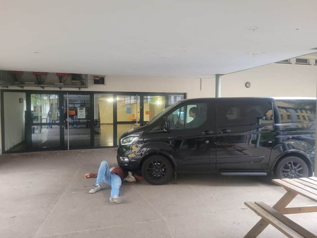 Eine Person liegt auf dem Boden, teilweise unter einem schwarzen Lieferwagen, der in der Nähe eines Gebäudeeingangs geparkt ist, und in der Nähe steht ein Picknicktisch.