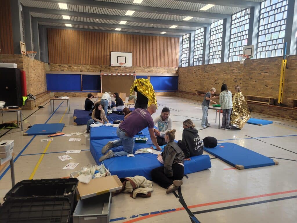 In einer Turnhalle üben Menschen auf Matten Erste-Hilfe-Maßnahmen, wobei sich einige um andere kümmern und Notfalldecken verwenden. Die Hilfsmittel liegen verstreut auf dem Boden.