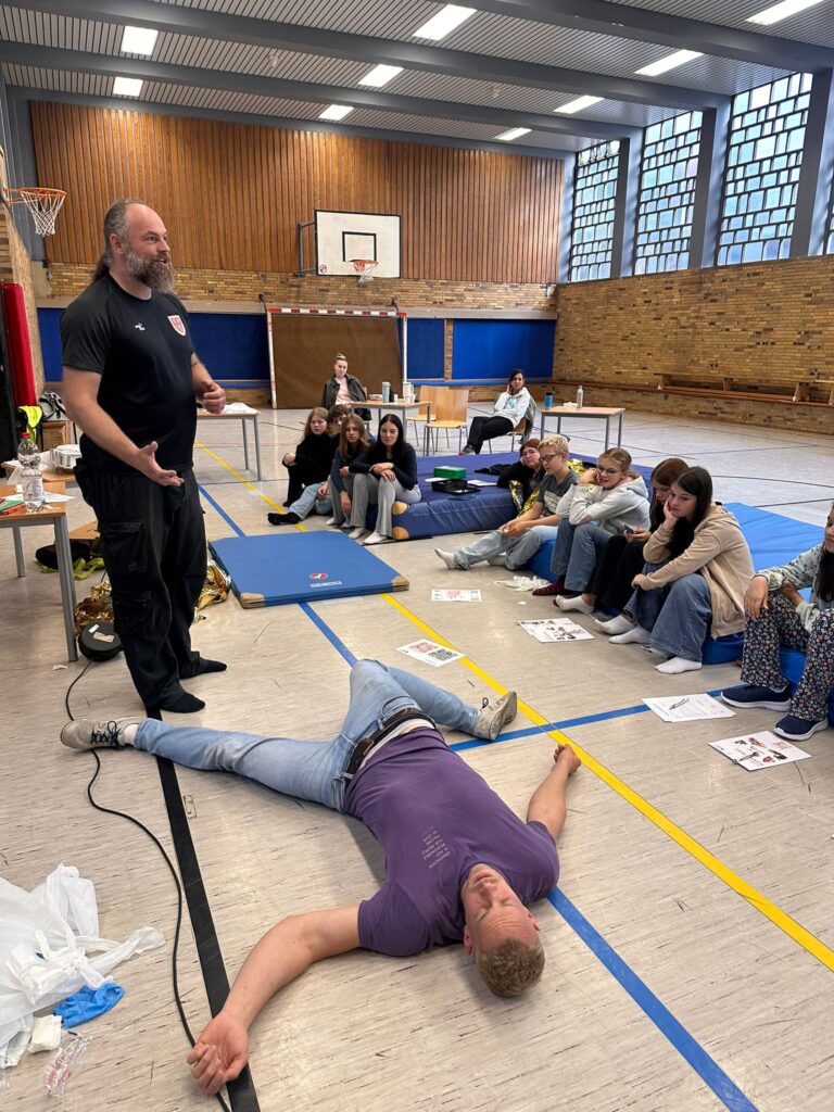 Ein Mann gibt einer Gruppe von sitzenden Personen in einer Turnhalle einen Erste-Hilfe-Kurs, während ein anderer Mann als Demonstrationsobjekt auf dem Boden liegt.