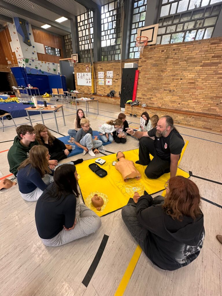 Eine Gruppe von Schülern sitzt im Kreis um einen Ausbilder und eine HLW-Puppe auf einer gelben Matte in einer Turnhalle und lernt Erste-Hilfe-Maßnahmen.