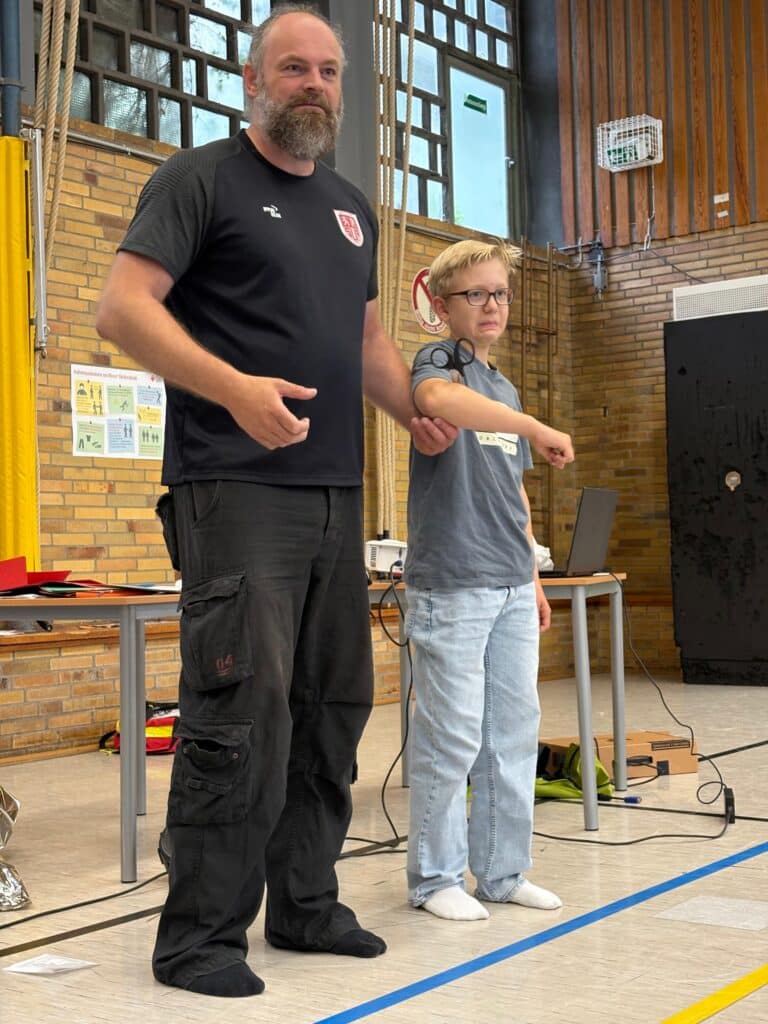 Ein bärtiger Mann in einem schwarzen Hemd demonstriert eine Technik, indem er einen Jungen in einer Sporthalle mit Backsteinwänden und Sportgeräten am Arm festhält.