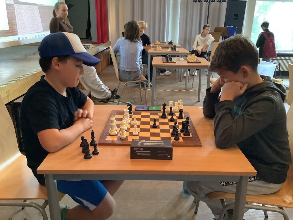Zwei Jungen sitzen sich bei einem Turnier gegenüber und spielen Schach, wobei im Hintergrund andere Spieler und Schachbretter zu sehen sind.
