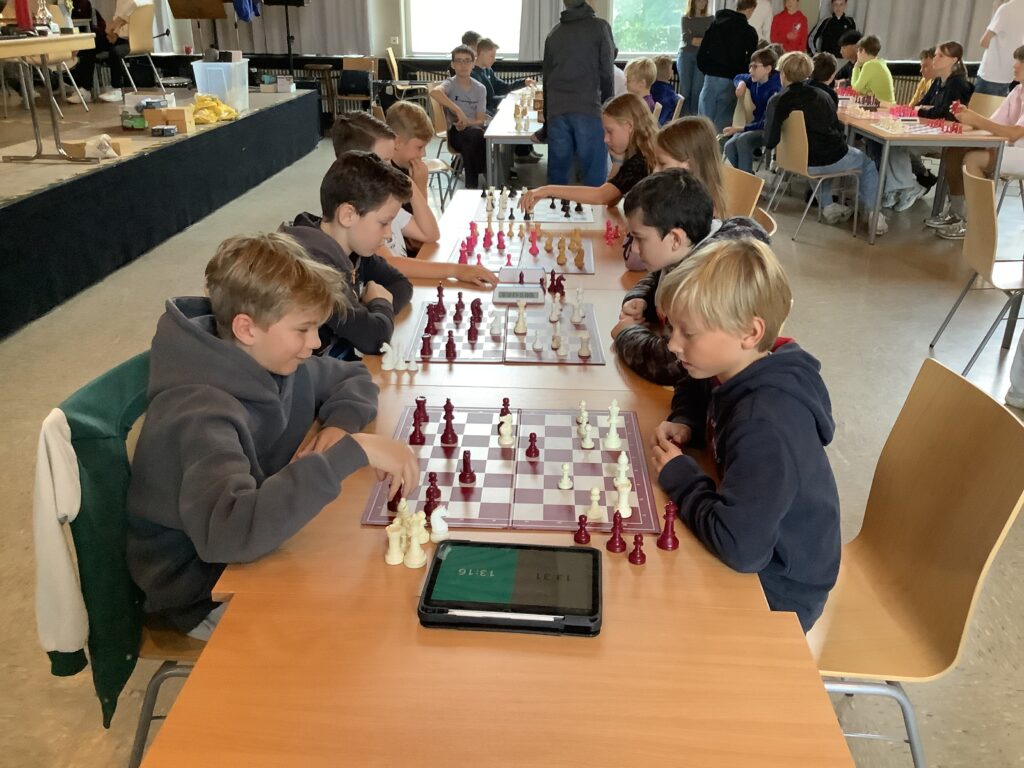Kinder sitzen sich an Tischen gegenüber und spielen Schach in einer Turnierumgebung. Mehrere Schachbretter und eine elektronische Schachuhr sind zu sehen.