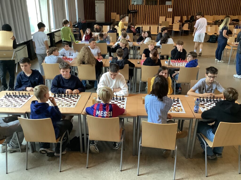 Eine Gruppe von Kindern und Jugendlichen spielt an mehreren Tischen in einem großen, hellen Raum Schach, wobei einige Leute zusehen und andere in der Nähe stehen oder sitzen.