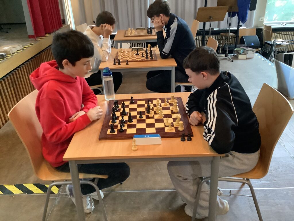 Vier Jungen spielen paarweise Schach an Holztischen in einem hellen Raum, konzentriert auf ihr Spiel. Ein Junge in einem roten Kapuzenpulli steht einem anderen in einer schwarzen Jacke gegenüber.