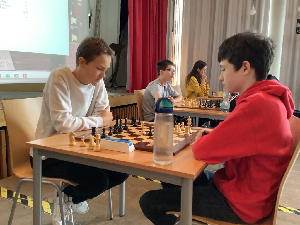 Zwei Jungen spielen Schach an einem Tisch, konzentriert auf das Spiel. Andere Schüler im Hintergrund spielen ebenfalls Schach. Eine Wasserflasche steht auf dem Tisch.