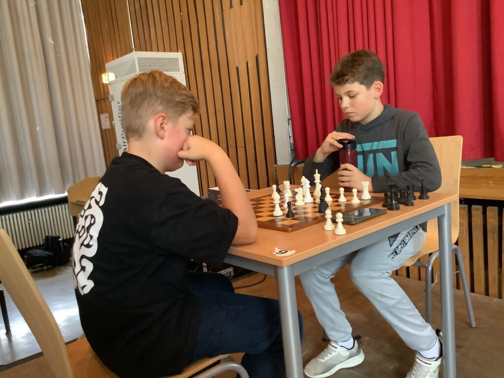 Zwei Jungen sitzen an einem Tisch und spielen in einem Haus Schach. Der eine denkt nach, der andere hält eine Flasche, und hinter ihnen ist ein roter Vorhang.