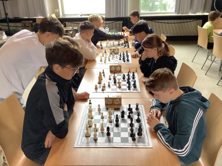 Mehrere Teenager sitzen sich an Tischen gegenüber und spielen konzentriert Schach. Zwischen jedem Paar sind Schachuhren aufgestellt.