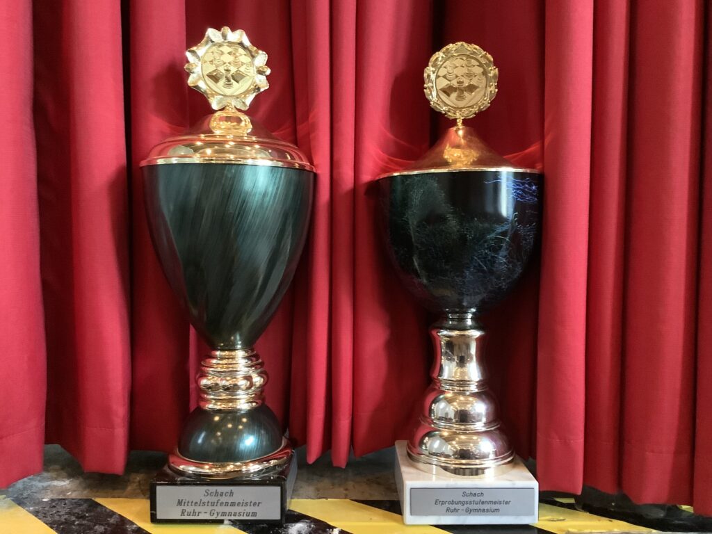 Zwei große goldene und grüne Trophäen sind vor roten Vorhängen ausgestellt, jede mit einer Plakette am Sockel und einem goldenen Schachemblem obenauf.