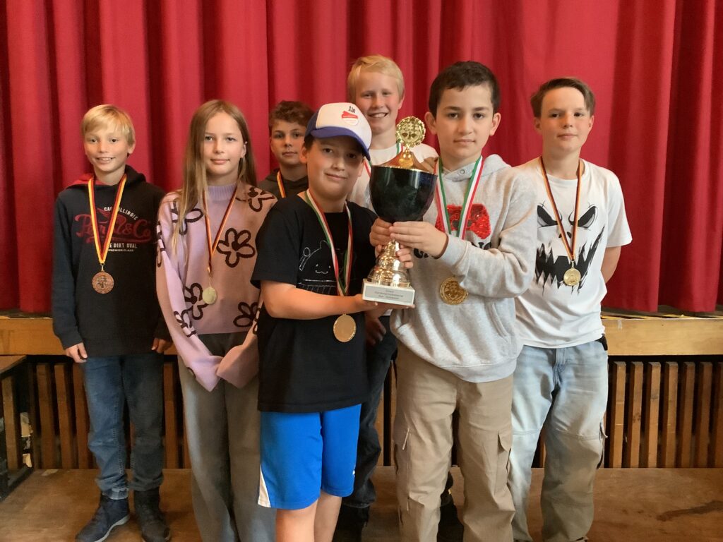 Sieben Kinder mit Medaillen stehen lächelnd vor einem roten Vorhang, zwei von ihnen halten gemeinsam einen großen Pokal.