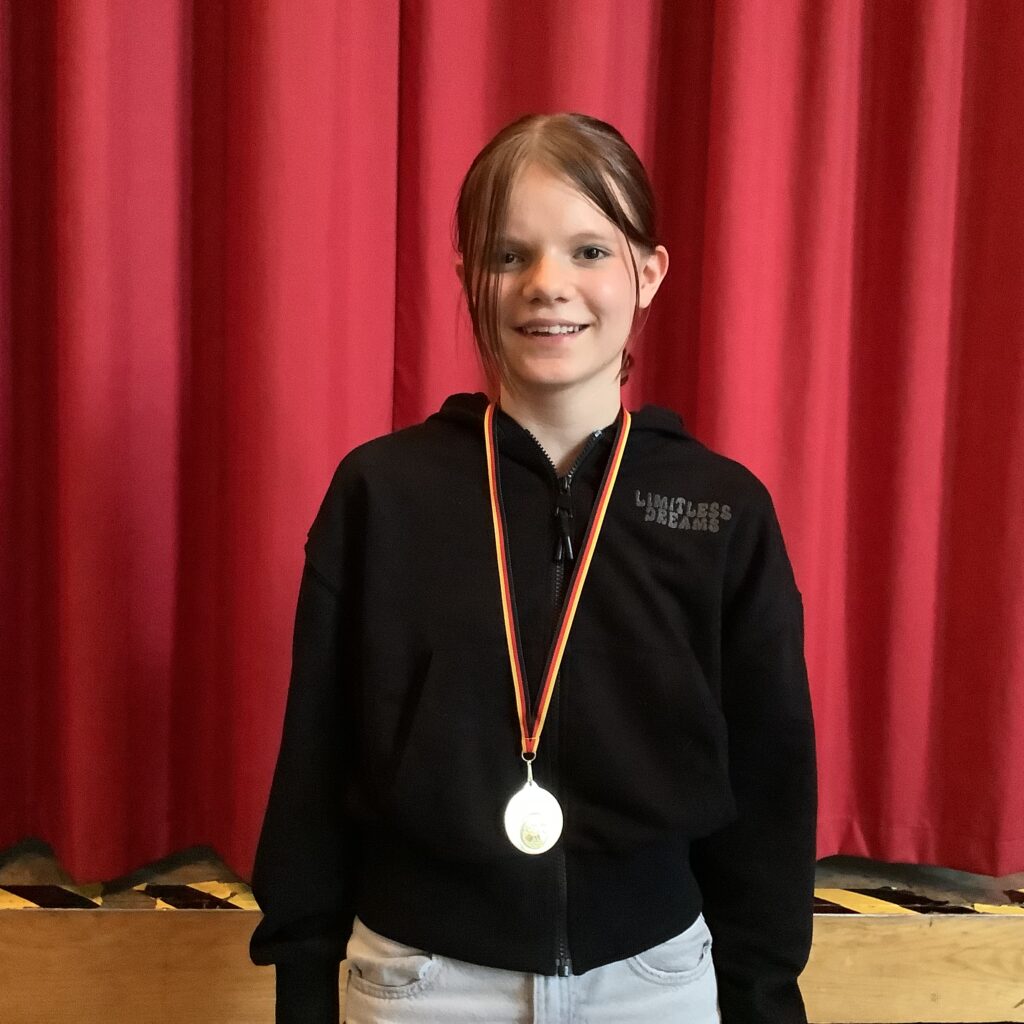 Ein lächelndes Mädchen mit einem schwarzen Kapuzenpulli und einer Medaille steht vor einem roten Vorhang.