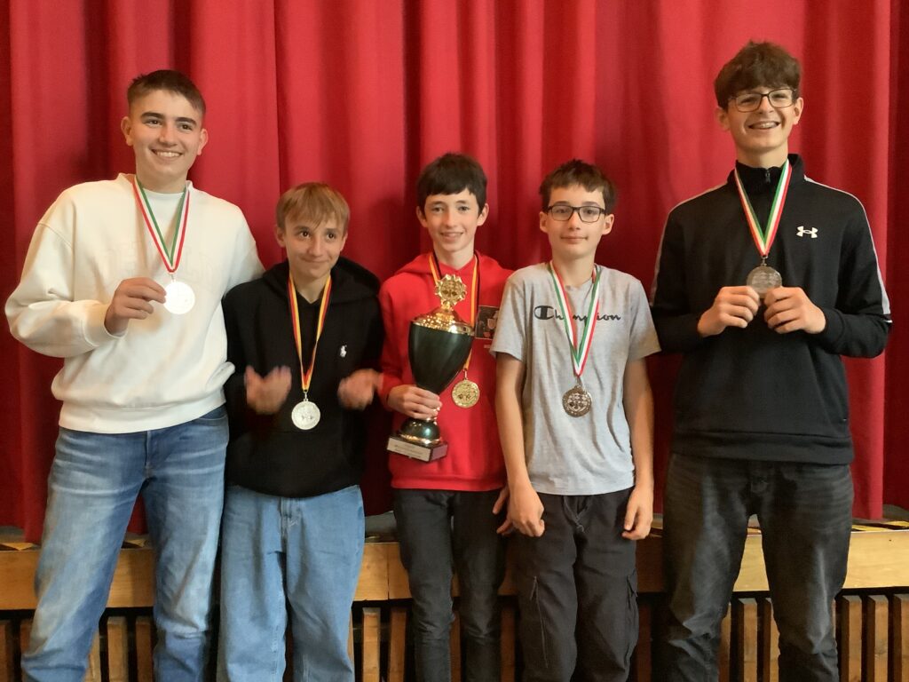 Fünf lächelnde Jungen stehen vor einem roten Vorhang, jeder trägt eine Medaille, während der Junge in der Mitte eine große Trophäe hält.