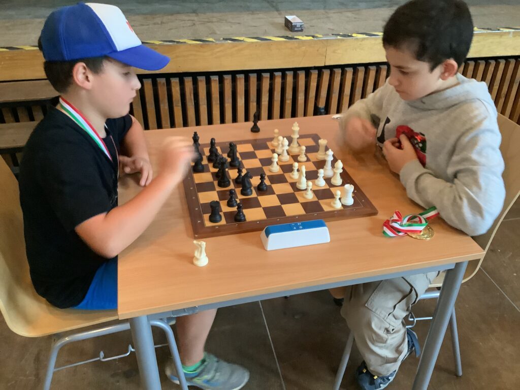 Zwei Jungen spielen eine Partie Schach an einem Holztisch. Beide tragen Medaillen, und eine Schachuhr liegt zwischen ihnen auf dem Tisch.