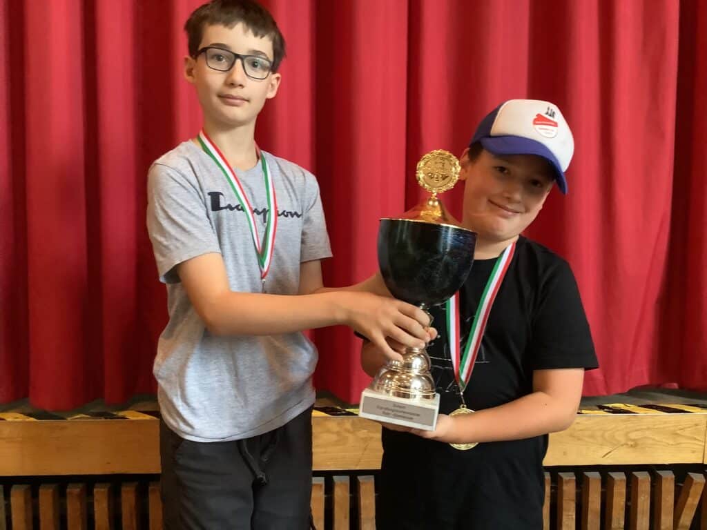 Zwei Jungen mit Medaillen halten gemeinsam einen Pokal vor einem roten Vorhang. Der eine trägt eine Brille und ein graues Hemd, der andere ein schwarzes Hemd und eine weiße Mütze.
