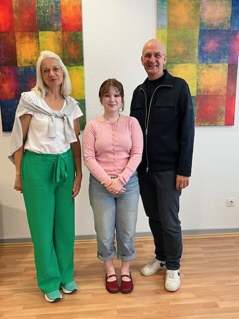 Drei Personen stehen in einem Innenraum auf einem Holzboden und posieren vor farbenfroher abstrakter Kunst. Die Gruppe besteht aus zwei Frauen und einem Mann, die alle in die Kamera lächeln.