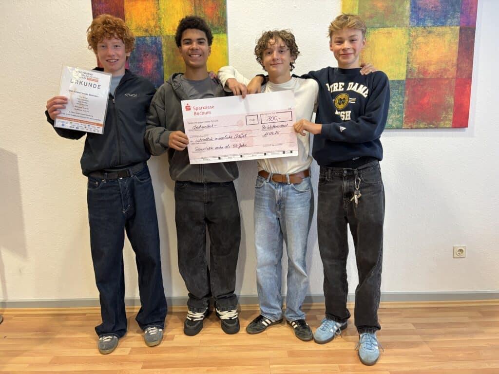 Vier lächelnde Teenager stehen in einem Raum; zwei halten einen großen Scheck und einer eine Urkunde in der Hand. Hinter ihnen hängt bunte abstrakte Kunst an der Wand.