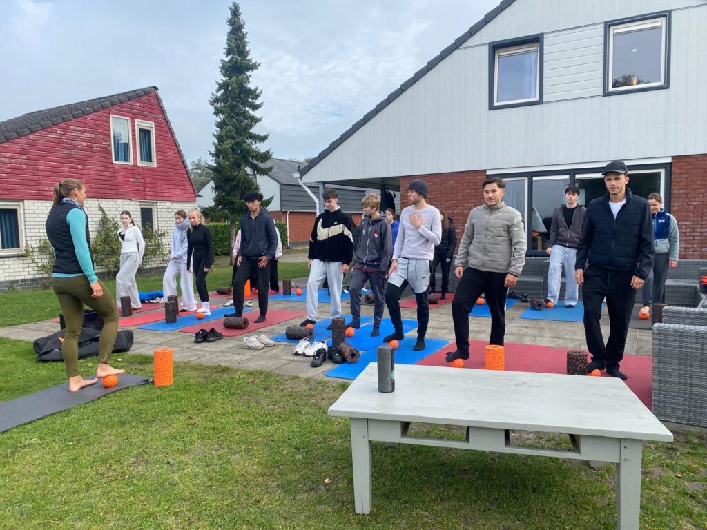 Eine Gruppe von Menschen steht auf Yogamatten im Freien und hört einem Lehrer zu. Sie sind leger gekleidet und scheinen sich auf eine Fitnessaktivität vorzubereiten.