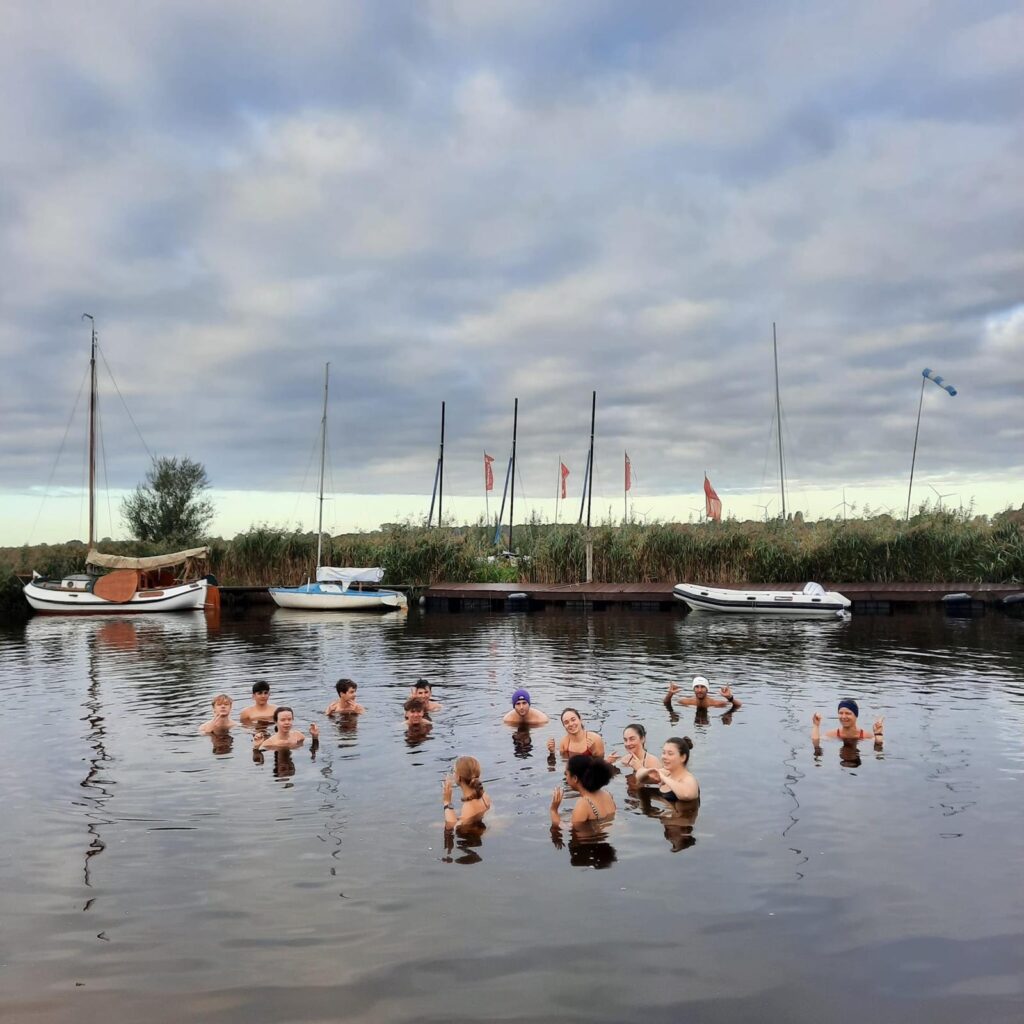 Eine Gruppe von Menschen schwimmt gemeinsam in einem ruhigen Fluss in der Nähe von vertäuten Booten und Schilf unter einem bewölkten Himmel. Einige halten Getränke in der Hand und alle scheinen sich zu amüsieren.