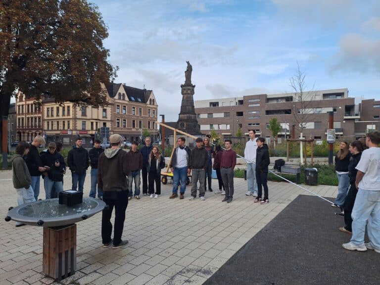 Eine Gruppe von Menschen steht auf einem Stadtplatz in der Nähe einer Statue, während eine Person ein Mikrofon über sie hält. Im Hintergrund sind Gebäude und Bäume zu sehen.