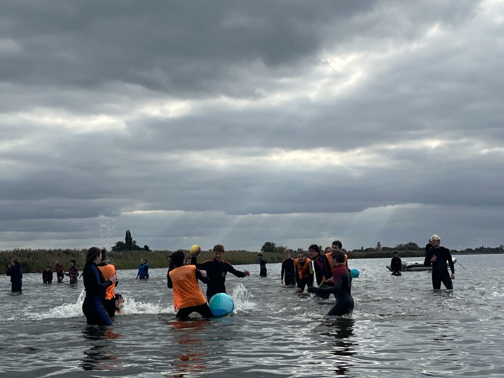 Eine Gruppe von Menschen in Neoprenanzügen und Schwimmwesten spielt mit großen blauen Bällen im flachen, bewölkten Wasser unter einem dramatischen, bedeckten Himmel.