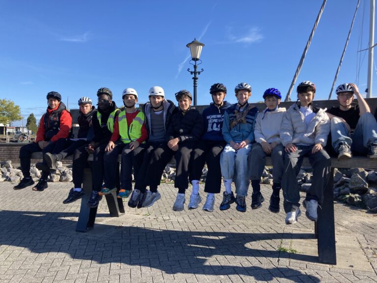 Zwölf Personen mit Helmen und Freizeitkleidung sitzen nebeneinander auf einer großen Holzbank im Freien unter blauem Himmel, mit Felsen und einem Laternenpfahl im Rücken.