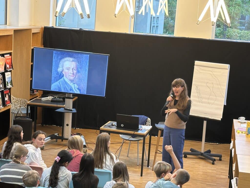 Eine Frau spricht zu einer Gruppe von Kindern in einer Bibliothek, während das Porträt eines älteren Mannes auf einem Bildschirm angezeigt wird und ein Flipchart neben ihr steht.