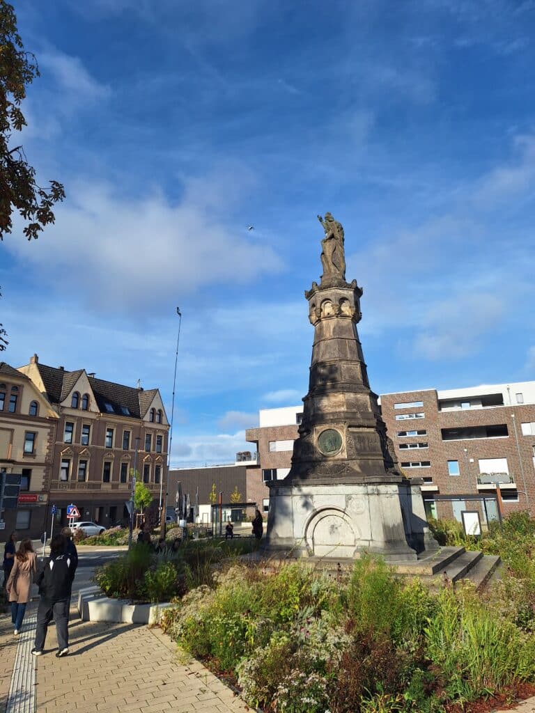 Ein hohes Steinmonument mit einer Statue auf der Spitze steht auf einem Stadtplatz, umgeben von Pflanzen, mit Menschen in der Nähe und Gebäuden im Hintergrund unter blauem Himmel.