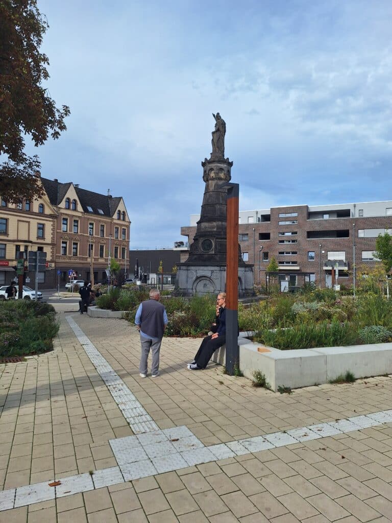Ein Stadtplatz mit einer großen Steinstatue in der Mitte, umgeben von Gebäuden, Pflanzen und zwei Menschen, die sich unter einem teilweise bewölkten Himmel auf Bänken unterhalten.