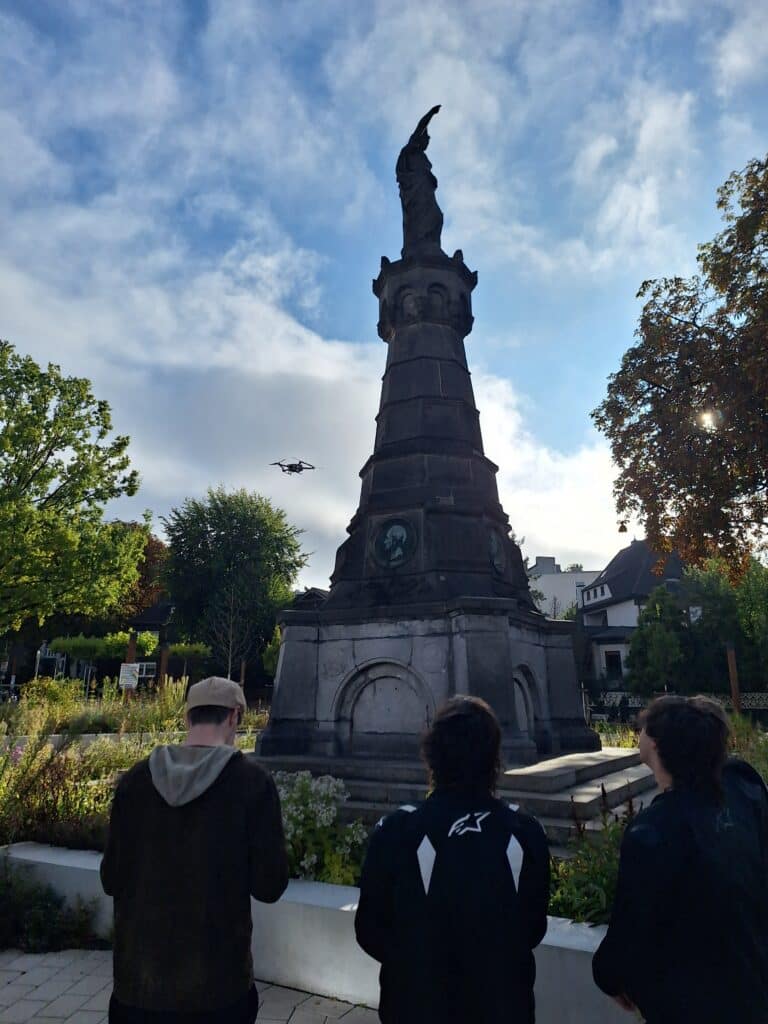 Drei Personen stehen vor einem hohen Steinmonument in einem Park, während eine Drohne bei teilweise bewölktem Himmel in der Nähe der Statue fliegt.