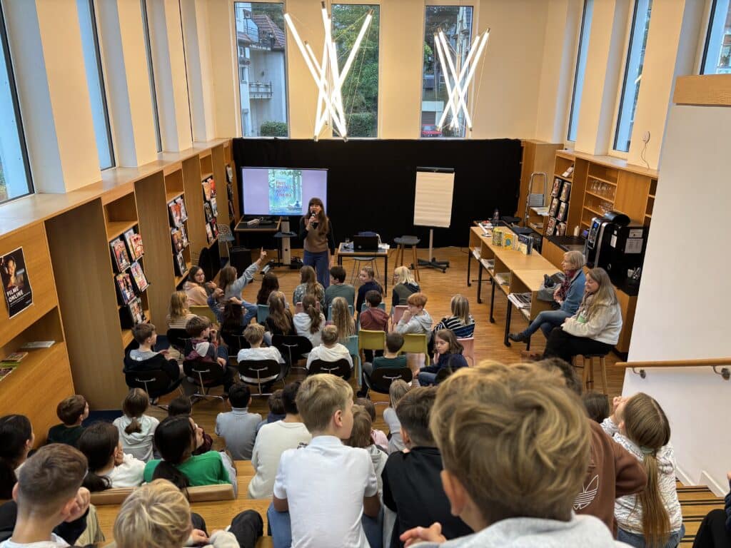 Eine Gruppe von Studenten sitzt in einer mehrstöckigen Bibliothek und sieht einer Frau zu, die vor einem Projektor und einem Flipchart spricht; an den Wänden stehen Bücherregale.