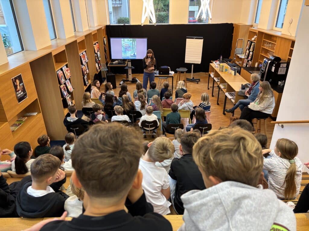 Eine Frau spricht zu einer Gruppe von Kindern, die in einem kleinen Bibliothekssaal sitzen, mit einer Leinwand, Büchern und einem Flipchart im vorderen Teil des Raums.