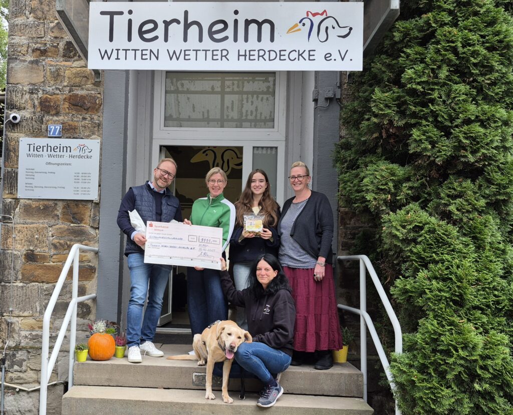 Fünf Personen stehen vor einem Tierheim, in der Hand einen großen Scheck, einen Hund und eine Spendenbox. Ein Hund sitzt mit einer Frau auf der Treppe.