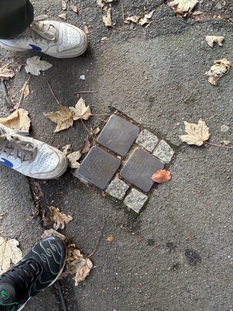 Drei Paar Schuhe stehen um vier Gedenktafeln aus Messing, die in den Gehweg eingelassen sind, umgeben von verstreuten Herbstblättern.