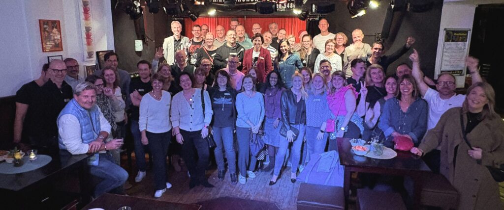 Eine große Gruppe lächelnder und miteinander posierender Menschen in einer warm beleuchteten Bar oder einem Club mit roten Vorhängen und kleinen Tischen im Vordergrund.