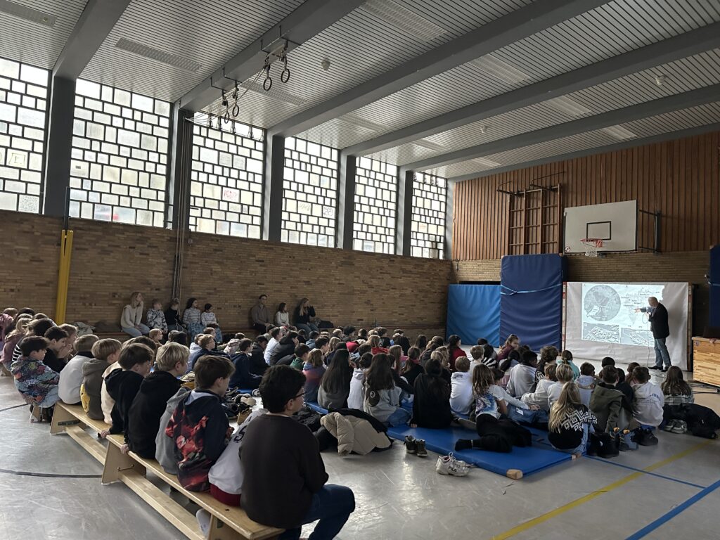 Eine große Gruppe von Schülern sitzt auf Bänken und Matten in einer Turnhalle und sieht zu, wie ein Redner Dias präsentiert, die auf eine Leinwand neben einem Basketballkorb projiziert werden.