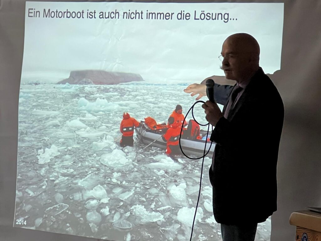 Ein Mann hält einen Vortrag und zeigt dabei auf ein projiziertes Bild von Menschen in orangefarbenen Anzügen auf einem Schlauchboot in eisigem Wasser. Auf dem Dia ist ein deutscher Text zu sehen.