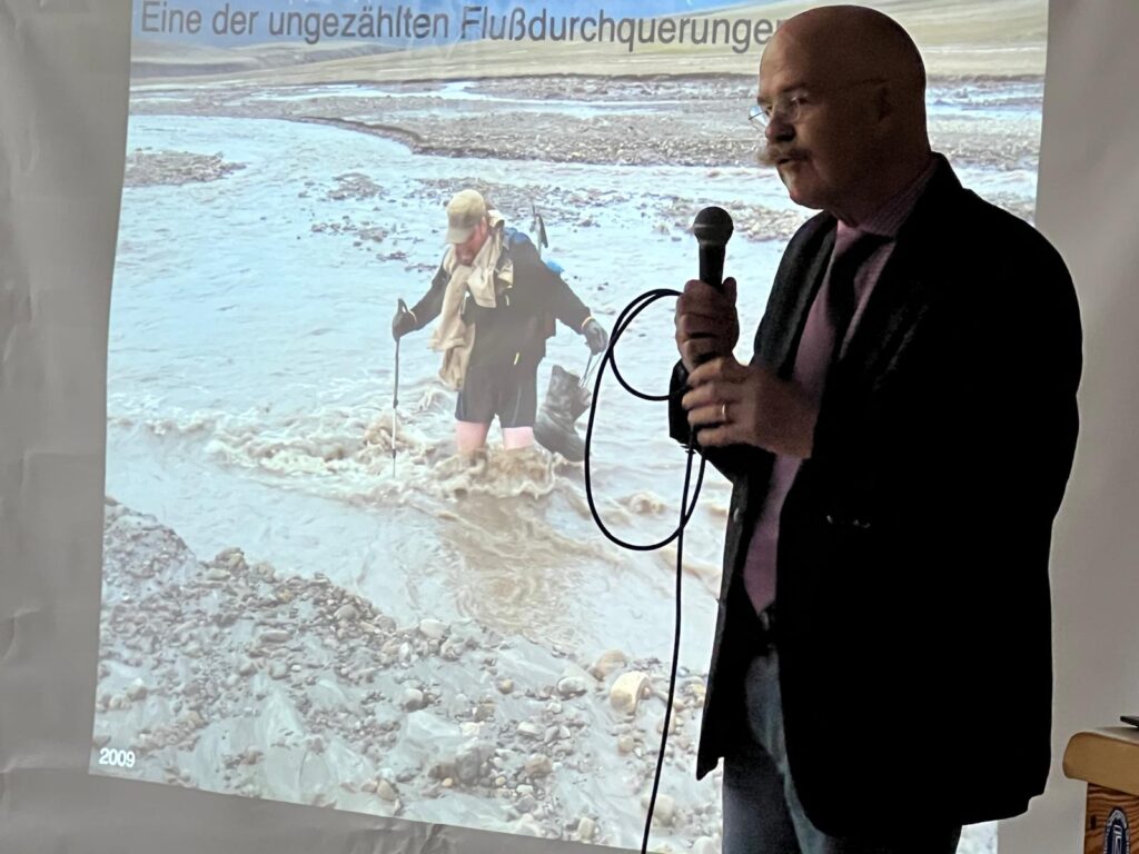 Ein Mann im Anzug hält ein Mikrofon, während er ein projiziertes Bild einer Person zeigt, die einen flachen, felsigen Fluss überquert. Am oberen Rand des Bildschirms ist ein deutscher Text zu sehen.