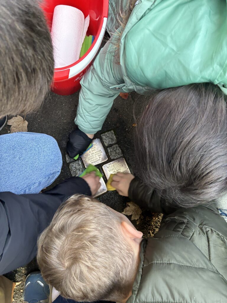 Vier Personen reinigen mit Tüchern und Schwämmen Messing-Gedenktafeln, die in einen Gehweg eingelassen sind. Ein roter Eimer mit Papierhandtüchern steht in der Nähe. Die Szene spielt sich im Freien ab.