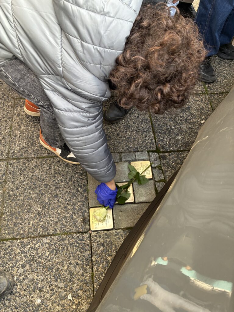 Eine Person mit lockigem Haar und grauer Jacke legt eine Rose auf eine in den Gehweg eingelassene Gedenktafel aus Messing und trägt einen blauen Handschuh.