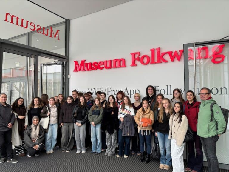 Eine Gruppe Jugendlicher und Erwachsener posiert gemeinsam vor dem Eingang des Museum Folkwang, vor großen Glastüren und einem leuchtend roten Schild.