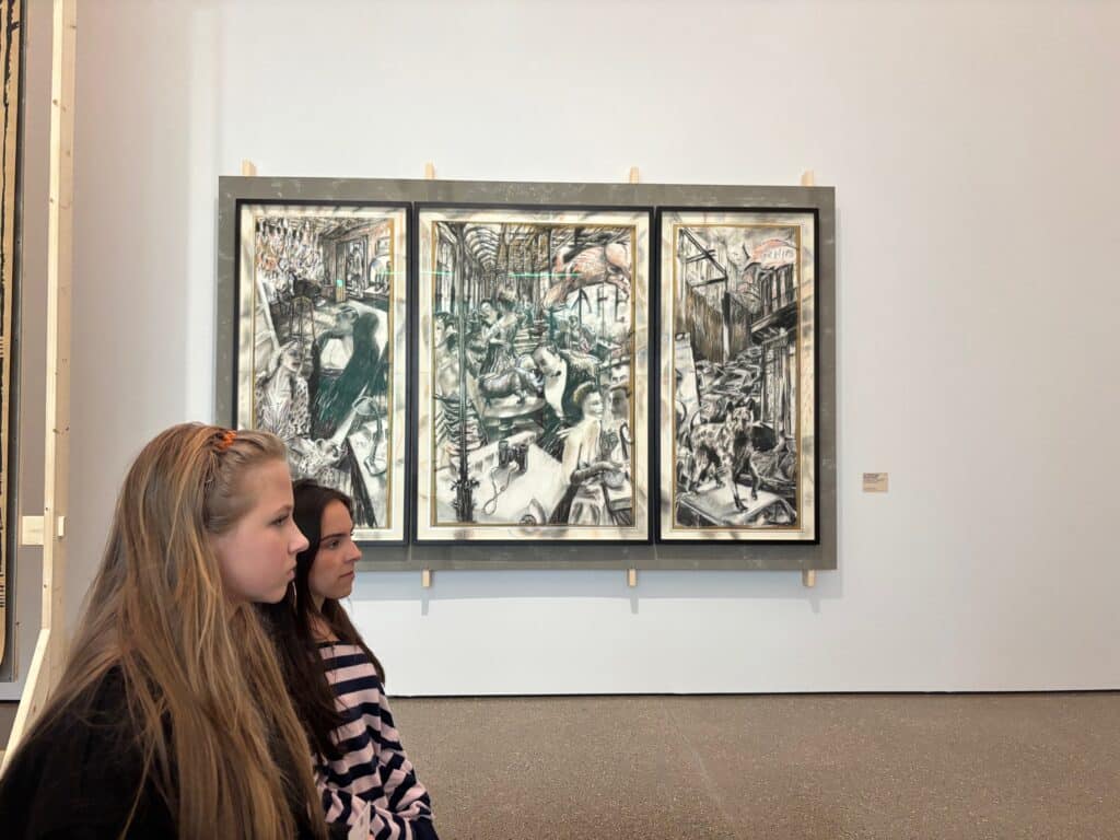 Zwei Mädchen sitzen vor einem großen, schwarz-weißen Triptychon an einer Galeriewand und betrachten das detailreiche und geschäftige Kunstwerk.