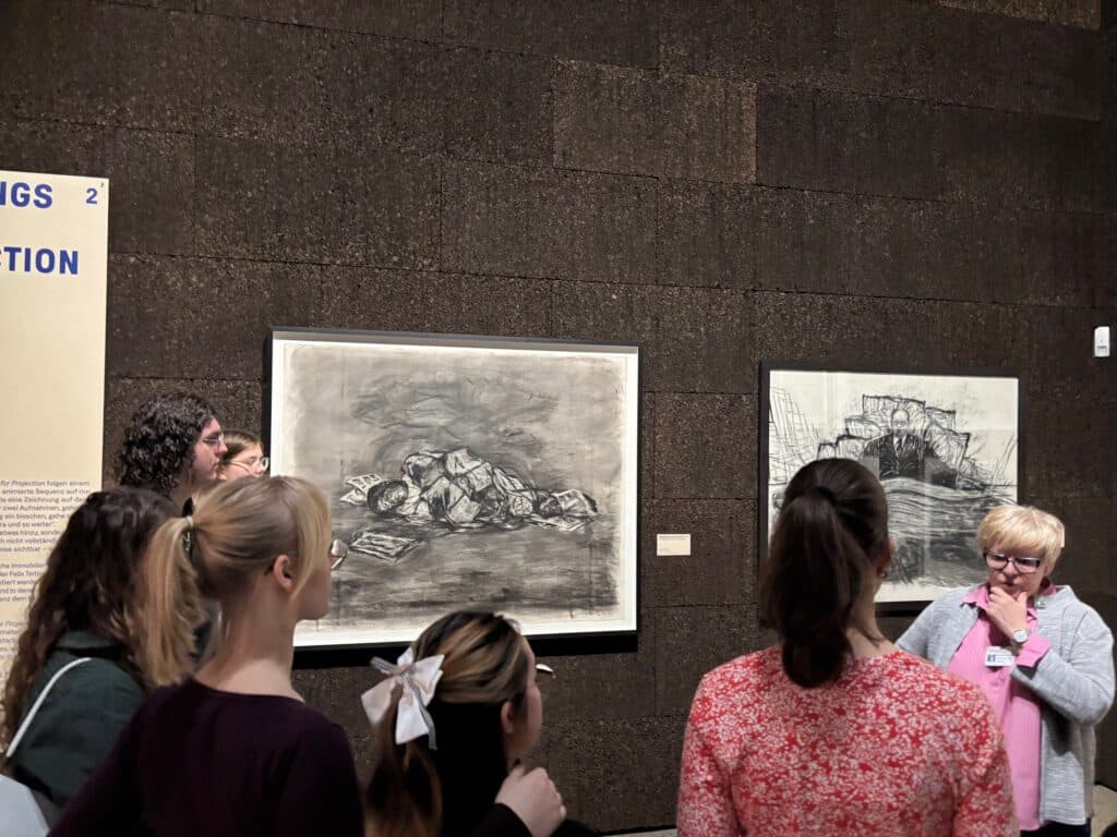 Eine Gruppe von Menschen hört einer Frau zu, die vor zwei großen Schwarz-Weiß-Zeichnungen an der Wand eines Museums spricht.