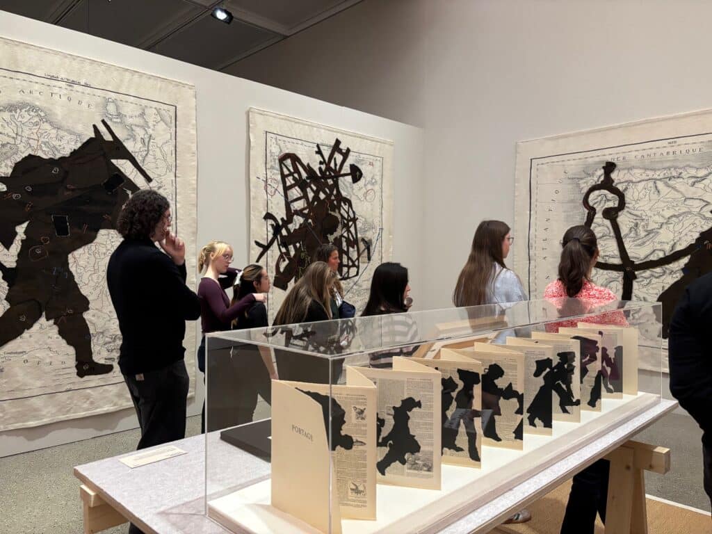 An den Wänden einer Galerie sind Kunstwerke mit Scherenschnitten und Landkarten zu sehen; in einer Vitrine im Vordergrund sind Bücher mit ausgeschnittenen Figuren ausgestellt.