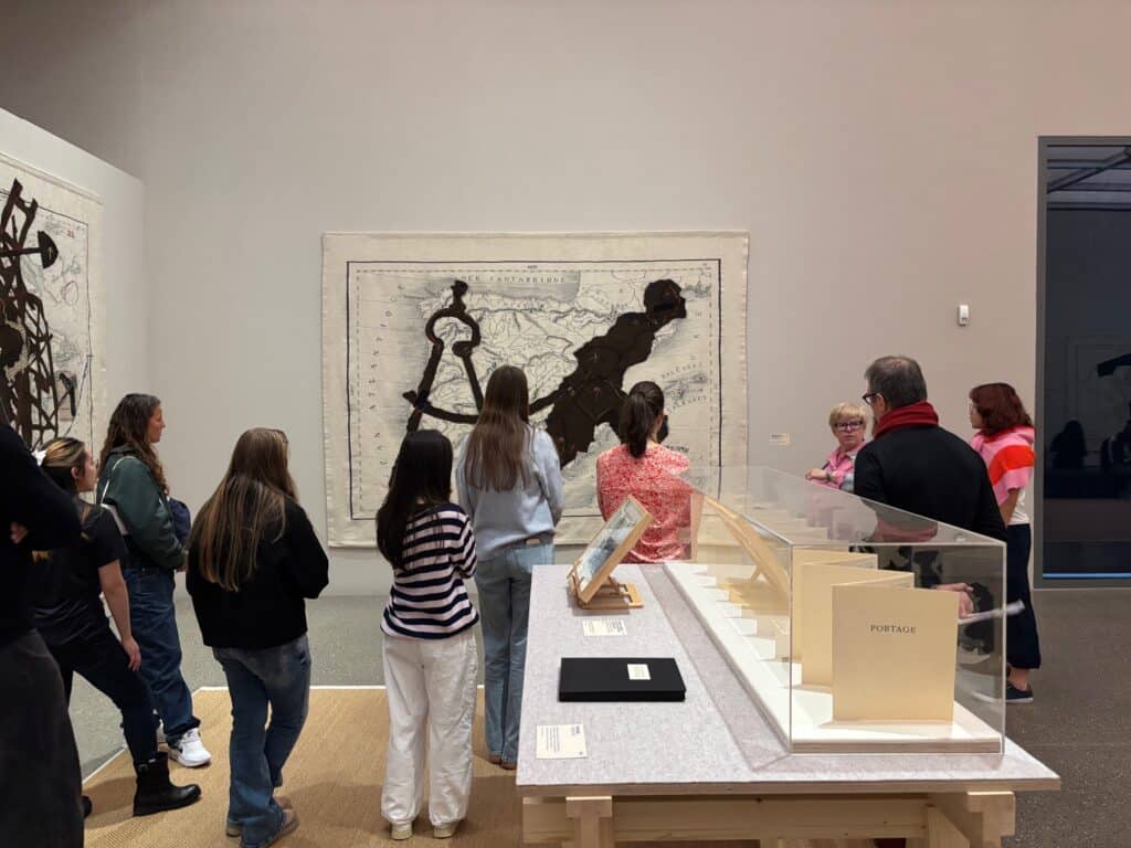 Eine Gruppe von Menschen steht in einer Kunstgalerie und hört einem Führer zu; an der Wand hängen Kunstwerke und im Vordergrund stehen Vitrinen mit Büchern.