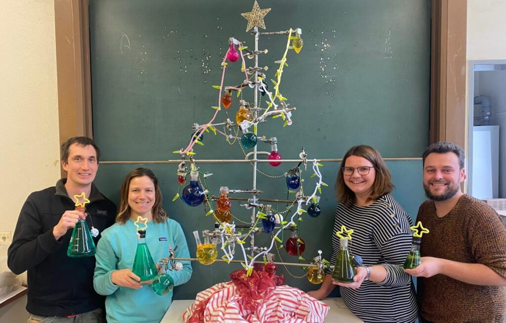 Vier Personen stehen lächelnd vor einem Weihnachtsbaum aus Laborgeräten und halten jeweils ein Fläschchen mit einem gelben Stern in der Hand. Der Baum steht vor einer Kreidetafel.