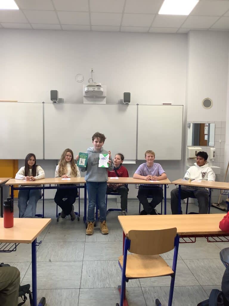 Ein Junge steht vor einem Klassenzimmer und hält zwei Zeichnungen hoch, während fünf andere Schüler an den Tischen hinter ihm sitzen und in die Kamera schauen.