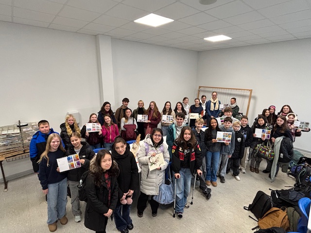 Eine Gruppe von Schülern und Erwachsenen steht in einem Klassenzimmer, einige halten bunte Papiere, alle lächeln und schauen in die Kamera. Um sie herum sind Mäntel und Taschen zu sehen.