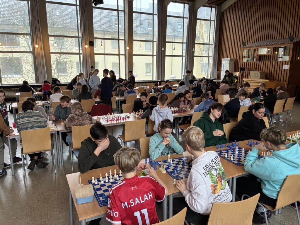Eine große Gruppe von Kindern spielt in einem hellen Raum mit hohen Fenstern Schach, sie sitzen an Tischen mit Schachbrettern und konzentrieren sich auf ihr Spiel.