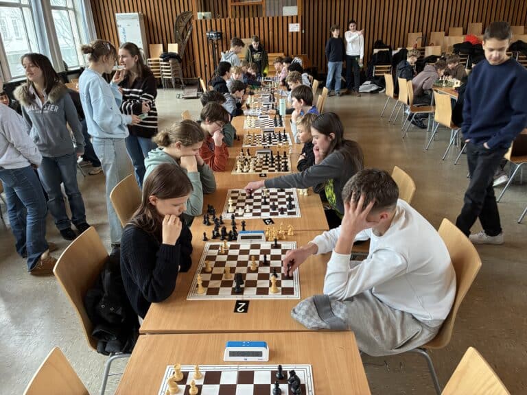 Die Schüler sitzen sich an langen Tischen gegenüber und konzentrieren sich auf das Schachspiel, während andere in einem hellen, modernen Raum stehen und zusehen.