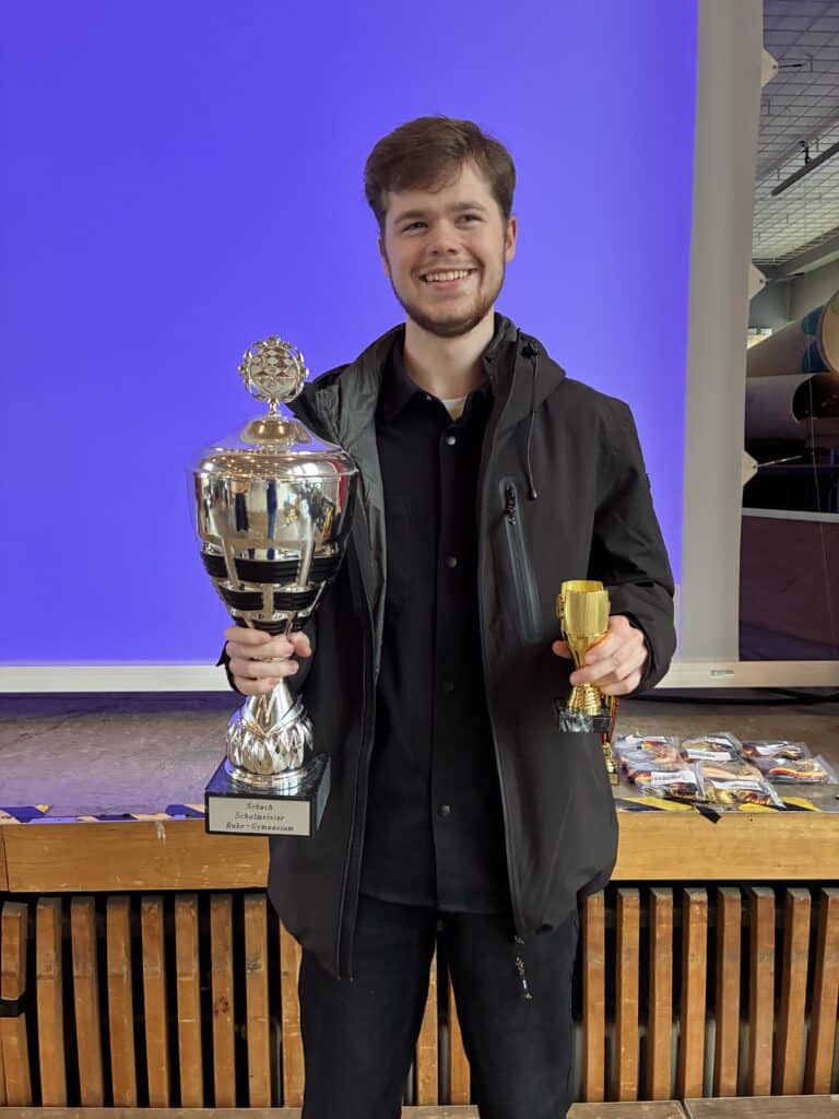 Ein lächelnder junger Mann in schwarzer Kleidung hält eine große silberne Trophäe in der einen und eine kleine goldene Trophäe in der anderen Hand und steht vor einem blauen Hintergrund.